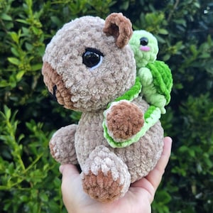 May include: A plush crochet capybara with light brown fur, dark brown accents on the nose and feet, and large black eyes. A small, light green crochet turtle with a darker green shell is attached to the back of the capybara, featuring black eyes.