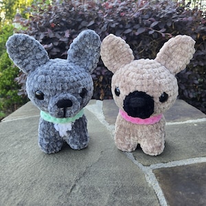 May include: Two handmade crochet French bulldog stuffed animals. One is gray with a green and white collar, the other is tan with a pink collar. Both have black eyes and noses, and large, floppy ears.