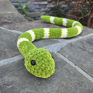 May include: A crocheted snake toy featuring a textured green body with repeating white and yellow striped bands. The plush snake has a rounded head with two small, smooth black plastic eyes positioned on the sides of its face.