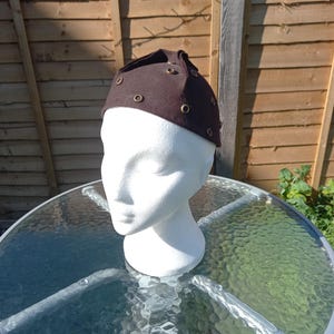 Può includere: Un cappello marrone scuro con una corona arrotondata e una serie di piccoli occhielli dorati. Il cappello è esposto su una testa di manichino bianca, sullo sfondo di una recinzione di legno e un tavolo di vetro.