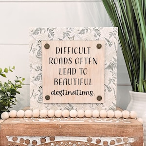 May include: Wooden sign with the quote "Difficult roads often lead to beautiful destinations." The sign is on a wooden box with a white and brown patterned design. A strand of wooden beads is on top of the box.