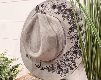 Puede incluir: Un sombrero fedora de fieltro gris con un diseño floral negro alrededor del borde. El sombrero está sentado en una caja de madera con un diseño estampado blanco y marrón.