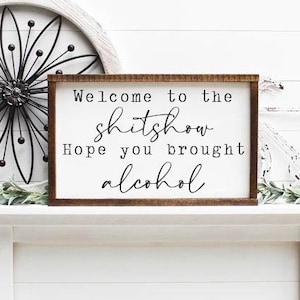 Può includere: Un cartello di legno con il testo "Welcome to the shitshow. Hope you brought alcohol." Il cartello è appoggiato su un camino bianco con un fiore di metallo bianco e una bicicletta di metallo bianco su entrambi i lati.