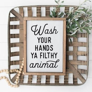May include: A wooden sign with the text "Wash your hands ya filthy animal" in black lettering. The sign is sitting in a woven basket with green branches.
