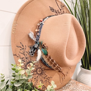Könnte beinhalten: Ein beigefarbener Fedora-Hut mit dekorativem Band und Federakzenten. Der Hut hat geätzte Blumendesigns und das Wort "leave" auf der Krempe. Er wird auf einer Holzoberfläche mit Grünzeug im Vordergrund präsentiert.