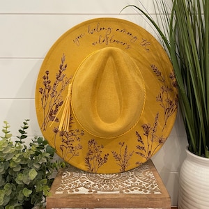 Puede incluir: Un sombrero fedora de fieltro amarillo mostaza con un borla marr&oacute;n y un dise&ntilde;o floral. El sombrero tiene el texto "you belong among the wildflowers" escrito en el ala.