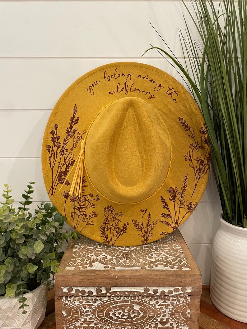 Puede incluir: Un sombrero fedora de fieltro amarillo mostaza con un borla marr&oacute;n y un dise&ntilde;o floral. El sombrero tiene el texto "you belong among the wildflowers" escrito en el ala.