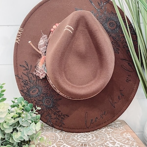 May include: A brown felt fedora hat with a black floral design and the text "leave her wild" on the brim.
