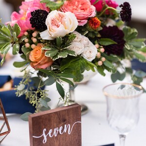 May include: A wooden table number sign with the word "seven" written in white calligraphy. The sign is sitting on a white tablecloth next to a bouquet of pink, white, and orange flowers.