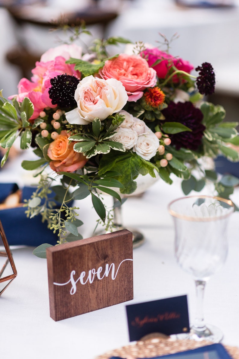 May include: A wooden table number sign with the word "seven" written in white calligraphy. The sign is sitting on a white tablecloth next to a bouquet of pink, white, and orange flowers.