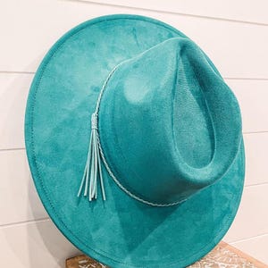 Puede incluir: Un sombrero fedora de gamuza azul turquesa con una borla de cuero azul claro y un ribete trenzado.