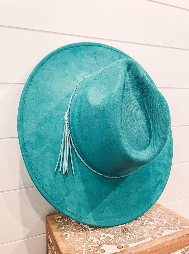 Puede incluir: Un sombrero fedora de gamuza azul turquesa con una borla de cuero azul claro y un ribete trenzado.