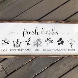 May include: A white wooden sign with the text "fresh herbs" in black cursive script.  The sign features black silhouettes of various herbs, including basil, rosemary, sage, dill, parsley, oregano, and thyme, with the name of each herb printed below.