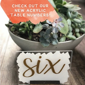 May include: Gold acrylic table number with the word "six" written in a calligraphy style font. The number is on a white background and is sitting on a wooden stand.