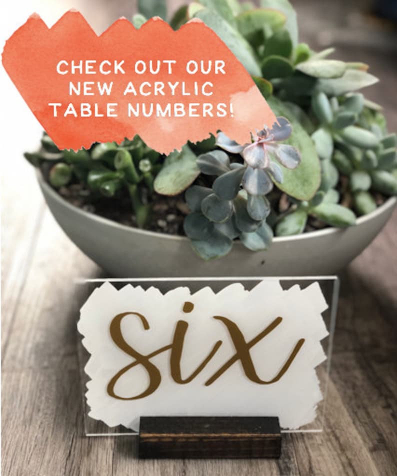 May include: Gold acrylic table number with the word "six" written in a calligraphy style font. The number is on a white background and is sitting on a wooden stand.