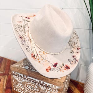 May include: A light beige cowboy hat with a floral design in shades of peach, burgundy, and green. The hat features a braided trim and a tassel. The hat is resting on a wooden box with carved details.