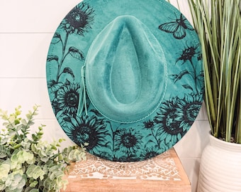 May include: A teal blue suede hat with a black sunflower and butterfly design. The hat is sitting on a wooden box with a white and brown patterned design.