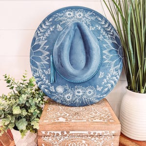 Puede incluir: Un sombrero azul estilo fedora con diseños florales y mariposas blancas. El sombrero tiene una borla a juego y se exhibe sobre una caja de madera con patrones tallados. Plantas decorativas en macetas blancas en el fondo.