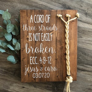 Könnte beinhalten: Ein Holzschild mit dem Text "A cord of three strands is not easily broken. ECC. 4:9-12 jesus & caro 03.07.20" und einem daran befestigten geflochtenen Seil.