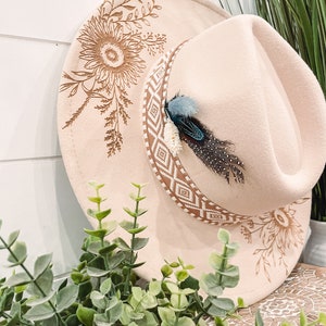 May include: A light beige cowboy hat with a sunflower design and a patterned band. The hat features a decorative feather accent. The brim is adorned with a sunflower and leaf design. The hat is a stylish accessory.
