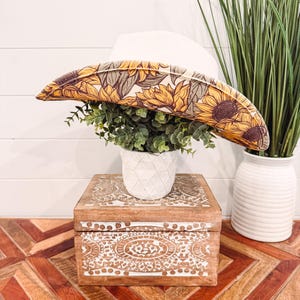 Puede incluir: Un sombrero de vaquero blanco con una banda de estampado de girasol. El sombrero está sentado en una caja de madera con un diseño intrincado. Una planta en maceta está delante del sombrero.