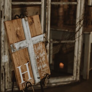 May include: A wooden sign with a white cross overlay and the letter "H" in white. The sign has a rustic, distressed look and includes the date "7.13.19". The sign is propped up on a black metal stand, suitable for weddings or home decor.