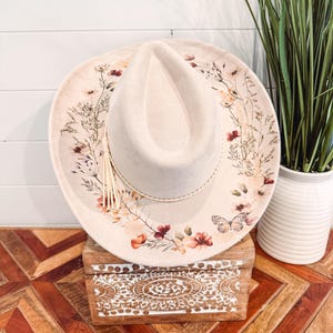May include: Cream-colored cowboy hat with a floral design on the brim, featuring red, orange, and yellow flowers, and a butterfly. The hat has a braided band and a tassel. The hat is sitting on a wooden box with a carved design.