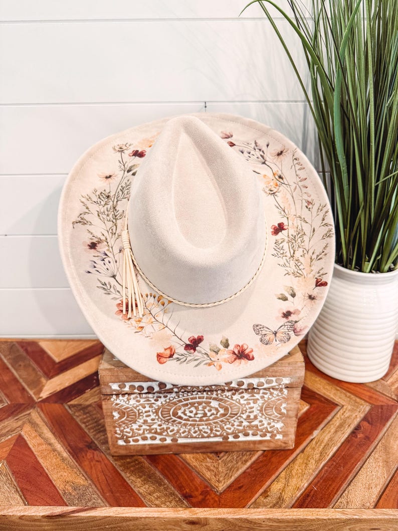 May include: Cream-colored cowboy hat with a floral design on the brim, featuring red, orange, and yellow flowers, and a butterfly. The hat has a braided band and a tassel. The hat is sitting on a wooden box with a carved design.