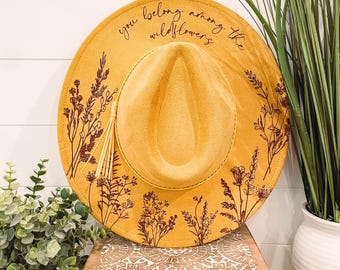 May include: A mustard yellow felt hat with a brown tassel and a floral design. The hat has the text "you belong among the wildflowers" written on the brim.