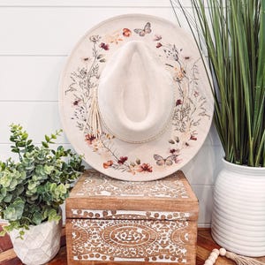 Op de afbeelding: Een cr&egrave;mekleurige hoed in fedora-stijl met een bloemen- en vlinderontwerp rond de rand. De hoed heeft een kwast en wordt getoond op een houten kist met gesneden details. De hoed heeft een diameter van ongeveer 35,5 cm.