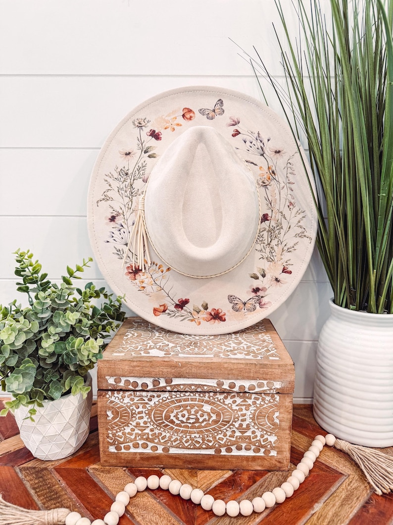 Op de afbeelding: Een cr&egrave;mekleurige hoed in fedora-stijl met een bloemen- en vlinderontwerp rond de rand. De hoed heeft een kwast en wordt getoond op een houten kist met gesneden details. De hoed heeft een diameter van ongeveer 35,5 cm.