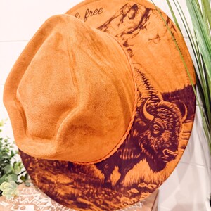 May include: A tan fedora hat with a wide brim. The brim has a dark brown illustration of a bison and mountains. The text "& free" is printed on the hat. The hat is made of a soft, suede-like material.