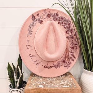 Puede incluir: Un sombrero de fieltro rosa con un dise&ntilde;o floral y el texto "wild child" en letras burdeos. El sombrero tiene una borla marr&oacute;n.