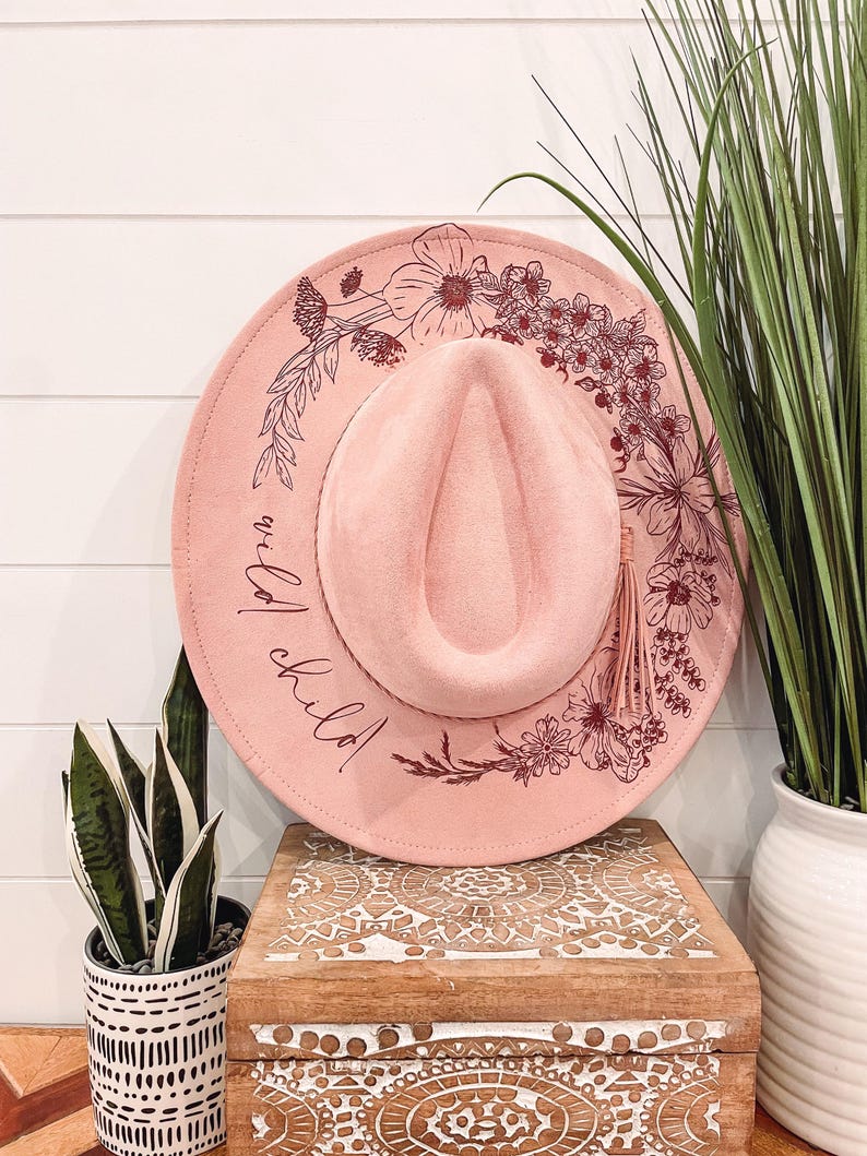 Puede incluir: Un sombrero de fieltro rosa con un dise&ntilde;o floral y el texto "wild child" en letras burdeos. El sombrero tiene una borla marr&oacute;n.