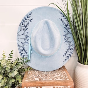 Puede incluir: Un sombrero de fieltro azul claro con un dise&ntilde;o floral azul. El sombrero tiene una borla azul claro y un ala ancha.