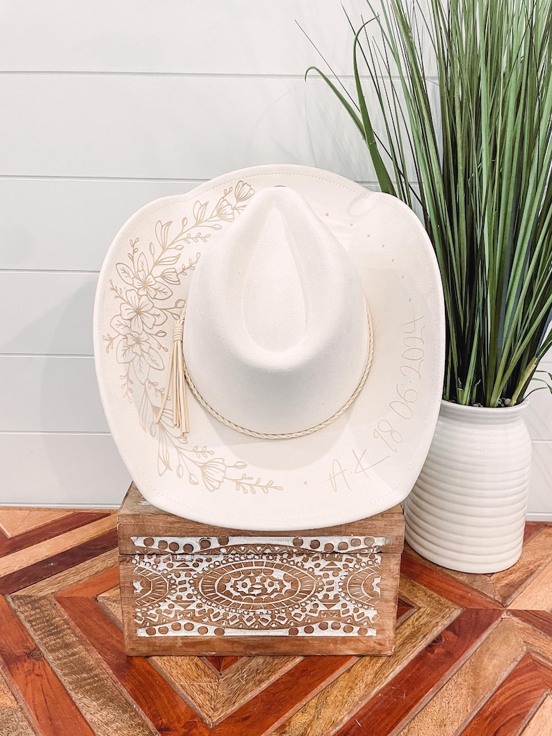 May include: A white cowboy hat with a floral design and the date "4.18.2024" written on the brim. The hat is sitting on a wooden box with an intricate design.