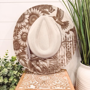 May include: A beige fedora-style hat with a wide brim featuring a brown floral and hummingbird design. The hat has a beige crown and a decorative tassel. The hat is displayed on a wooden box.