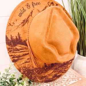May include: A tan fedora hat with a wide brim. The brim features a mountain and forest landscape design with the words "wild & free". The hat has a leather cord and tassel.