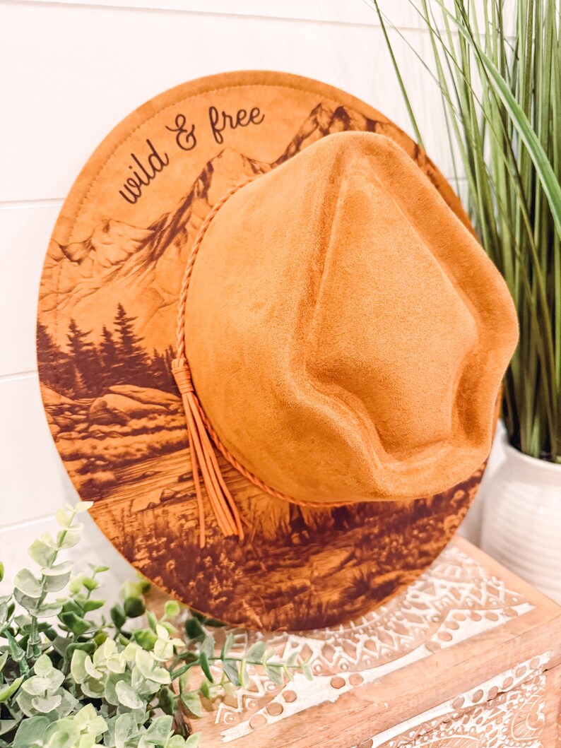 May include: A tan fedora hat with a wide brim. The brim features a mountain and forest landscape design with the words "wild & free". The hat has a leather cord and tassel.