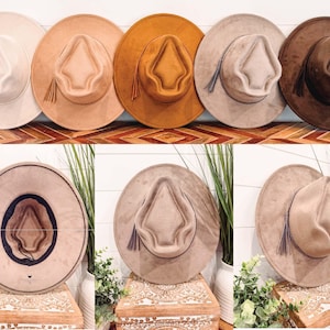 Op de afbeelding: Een collectie breedgerande hoeden in neutrale kleuren, waaronder crème, beige en bruin. Elke hoed heeft een gestructureerde kroon, een decoratieve kwast en een brede rand. De hoeden worden getoond op een houten oppervlak en tegen een witte achtergrond.