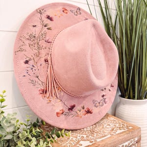 May include: A pink felt fedora hat with a floral and butterfly print. The hat has a brown leather band with a tassel.