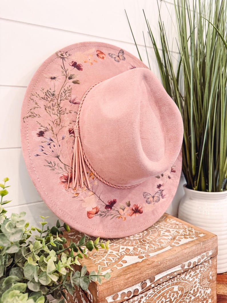 May include: A pink felt fedora hat with a floral and butterfly print. The hat has a brown leather band with a tassel.