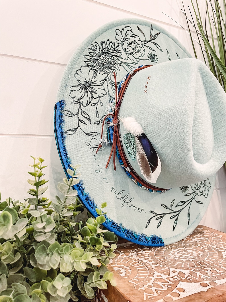 May include: A light blue felt hat with a floral design and the text "wildflower" on the brim. The hat has a blue and white trim and a brown leather band with a feather accent.