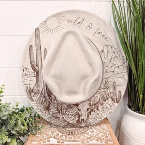 Op de afbeelding: Een beige fedora hoed met een woestijnontwerp. De hoed heeft een zon, cactussen, bergen en een schedel met de woorden "wild & free". De hoed wordt getoond op een houten kist.