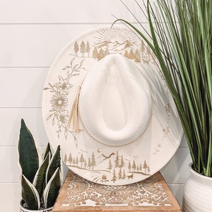 May include: White wide-brimmed hat with a gold mountain and forest scene design. The hat is sitting on a wooden box with a carved design.