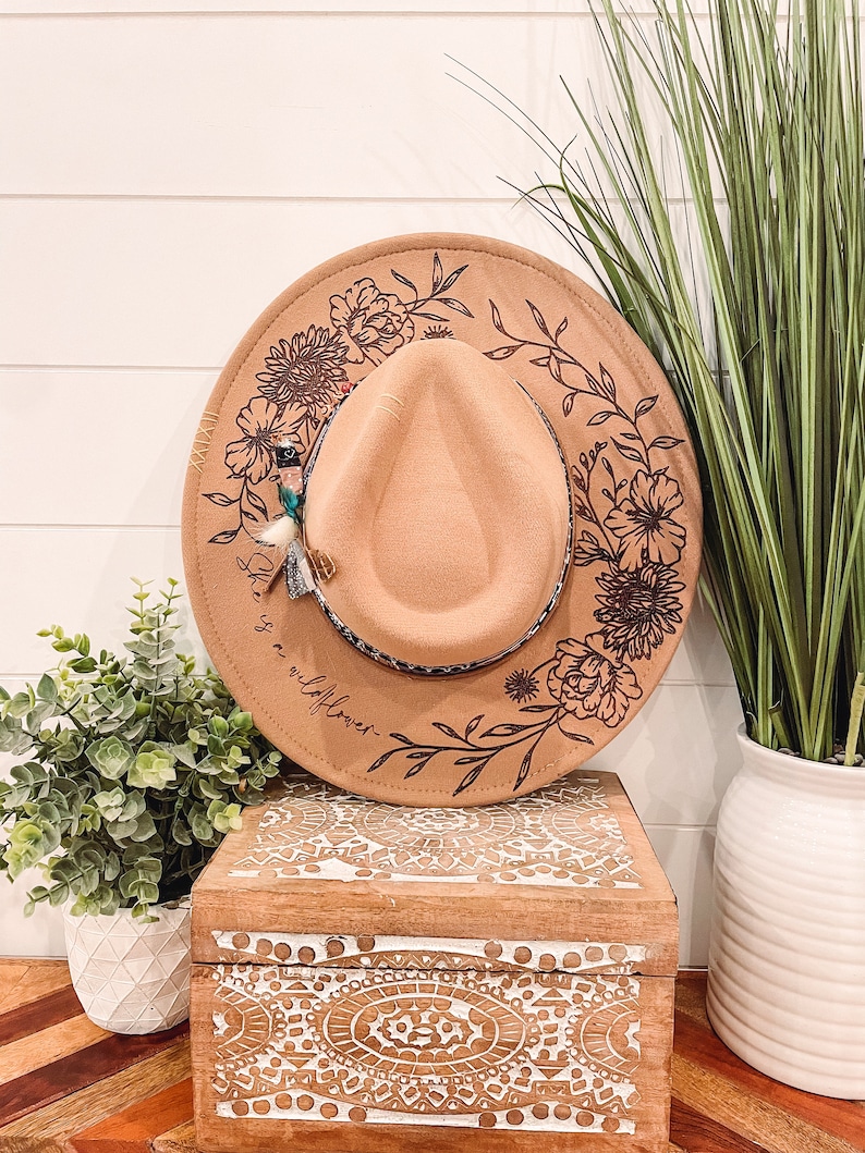 May include: A brown felt hat with a floral design and the text "Be a wildflower" on the brim. The hat is sitting on a wooden box with a white and brown patterned design.
