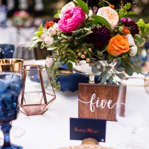 May include: A table setting with blue glassware, gold accents, and a wooden table number with the word "five" written in white. The centerpiece is a bouquet of pink, orange, and purple flowers.