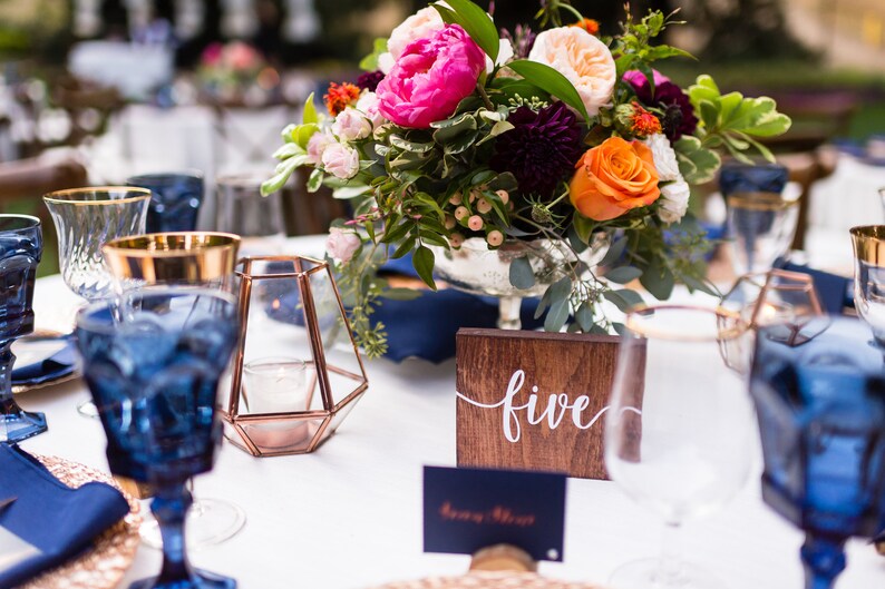 May include: A table setting with blue glassware, gold accents, and a wooden table number with the word "five" written in white. The centerpiece is a bouquet of pink, orange, and purple flowers.