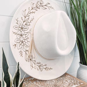 May include: A white fedora hat with a gold floral design. The hat has a wide brim and a small crown.