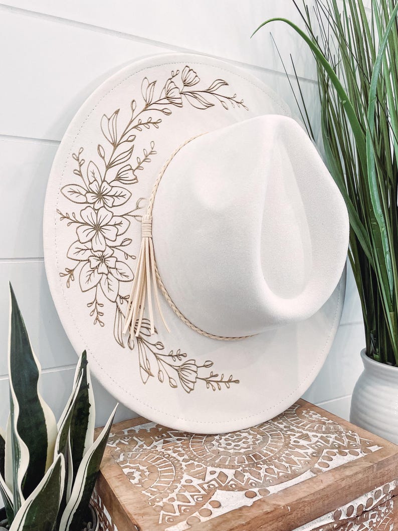 May include: A white fedora hat with a gold floral design. The hat has a wide brim and a small crown.